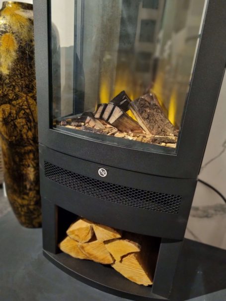 Modern glass-fronted fireplace with logs and warm lighting, situated in a sleek setting.