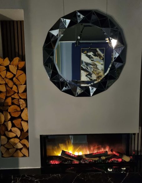 A modern fireplace with a circular mirror above and neatly stacked logs on the side